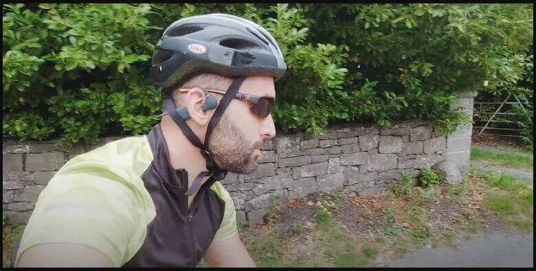 are bone conduction headphones safe for hearing? - a man riding a bicycle in full cycling gear; cycling helmet, sunglasses, and wearing an open-ear bone conduction headphone.